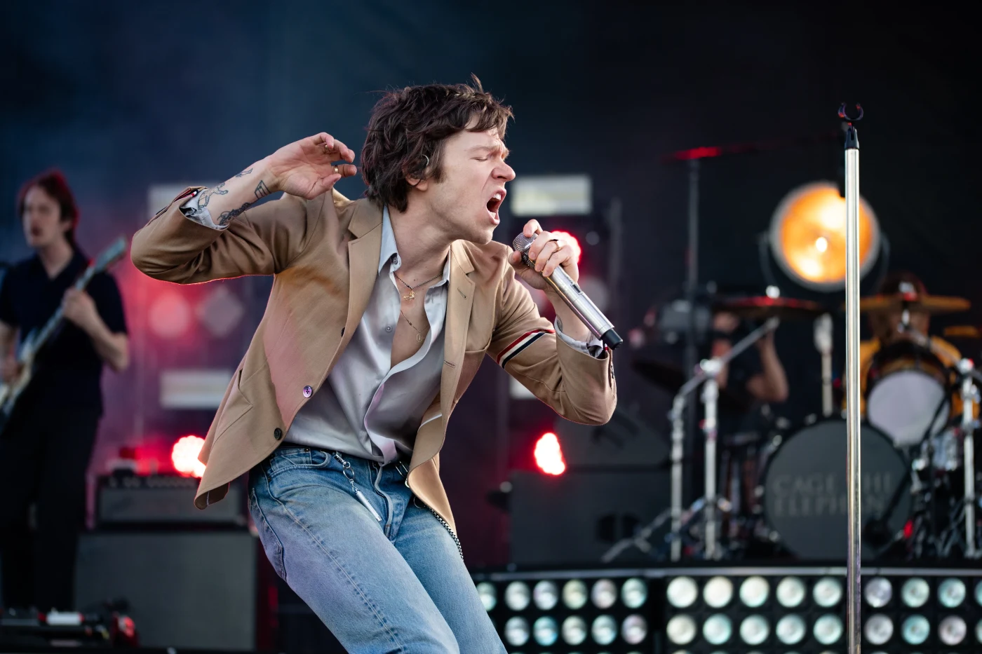 Rock frontman in a tan jacket singing mid-movement on stage